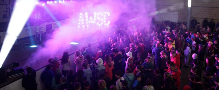 A disco with pink light at the All Wales Scout Camp. There is an AWSC logo lit up and lots of people dancing.